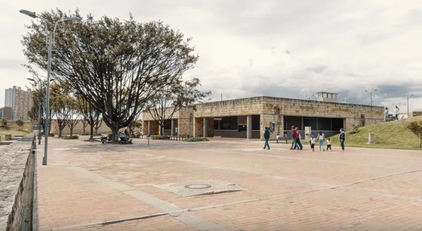 Plaza amplia en ladrillo al aire libre, con un edificio bajo de piedra y ventanales oscuros al fondo. A la izquierda, un árbol grande brinda sombra junto a bancas y faroles, mientras varias personas caminan por el espacio abierto en un entorno urbano con cielo nublado.
