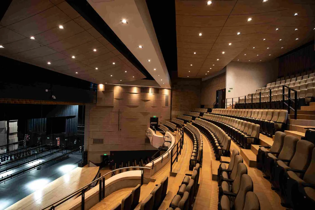 Interior de un teatro moderno con graderías semicirculares de asientos acolchados que miran hacia un escenario iluminado. El recinto tiene paredes en tonos cálidos, barandas negras y un techo con múltiples luces empotradas que iluminan el espacio.