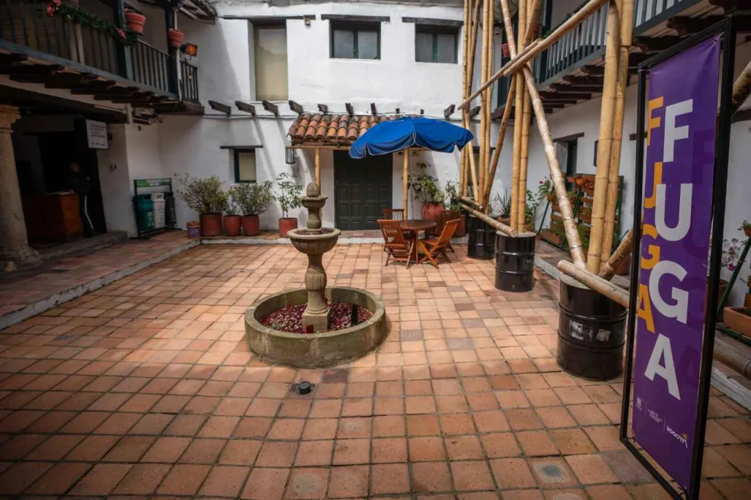 Patio interior de una casa colonial con piso de baldosas de barro, una fuente de piedra en el centro y mesas de madera con sombrilla azul. Alrededor hay balcones con barandas negras, plantas en macetas y una estructura de guadua junto a un cartel morado que dice “FUGA”.