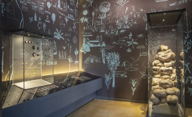  Interior de una sala de museo con iluminación tenue. En vitrinas de vidrio se exhiben objetos antiguos, como piezas metálicas y una estructura de piedras apiladas. Las paredes oscuras están decoradas con ilustraciones dibujadas en líneas blancas que representan paisajes, plantas, animales y escenas culturales, creando un ambiente histórico y educativo.