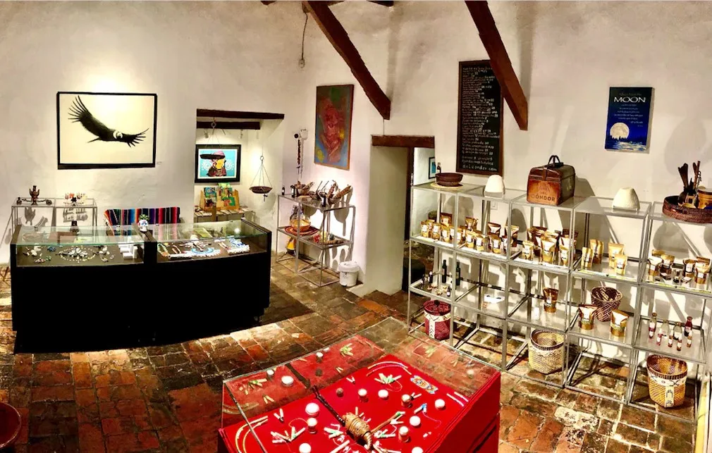 Interior de una tienda artesanal con piso de ladrillo y paredes blancas, decorada con vigas de madera en el techo. En el centro y a la izquierda hay vitrinas de vidrio que exhiben joyería y pequeños objetos hechos a mano. Al fondo, estanterías metálicas muestran frascos, productos naturales y canastos tejidos.