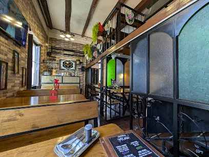 Interior de un restaurante de estilo rústico y acogedor, con paredes de ladrillo a la vista y vigas de madera en el techo. El espacio cuenta con mesas y bancas de madera, una barra al fondo y una estructura de dos niveles con barandas metálicas.