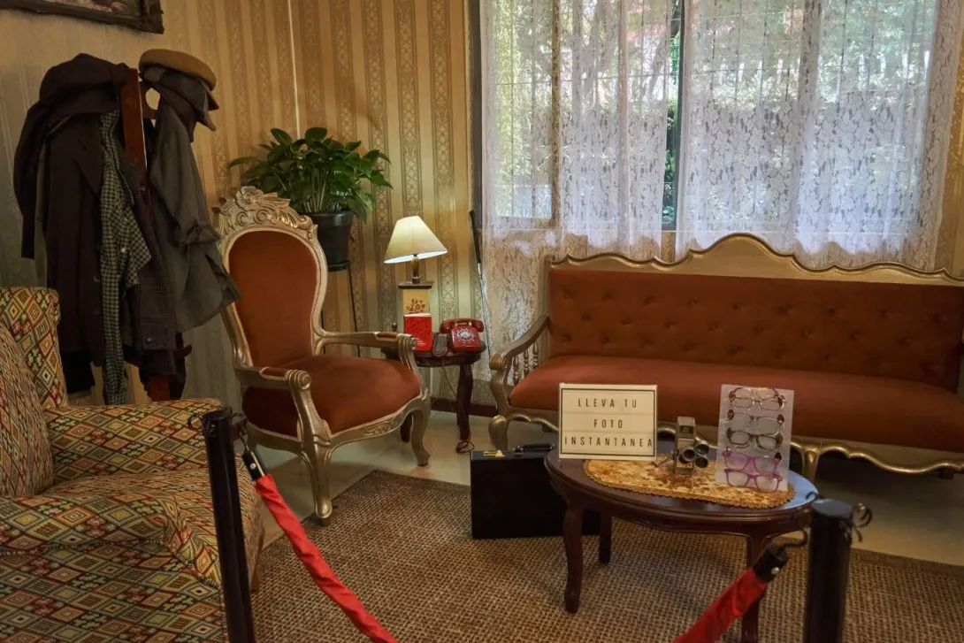 La imagen muestra un acogedor espacio interior de estilo vintage, con muebles clásicos como un sofá y sillones tapizados en tonos cálidos. Sobre una mesa se observan objetos antiguos, como un teléfono rojo y una cámara, junto a un letrero que invita a tomarse una foto.