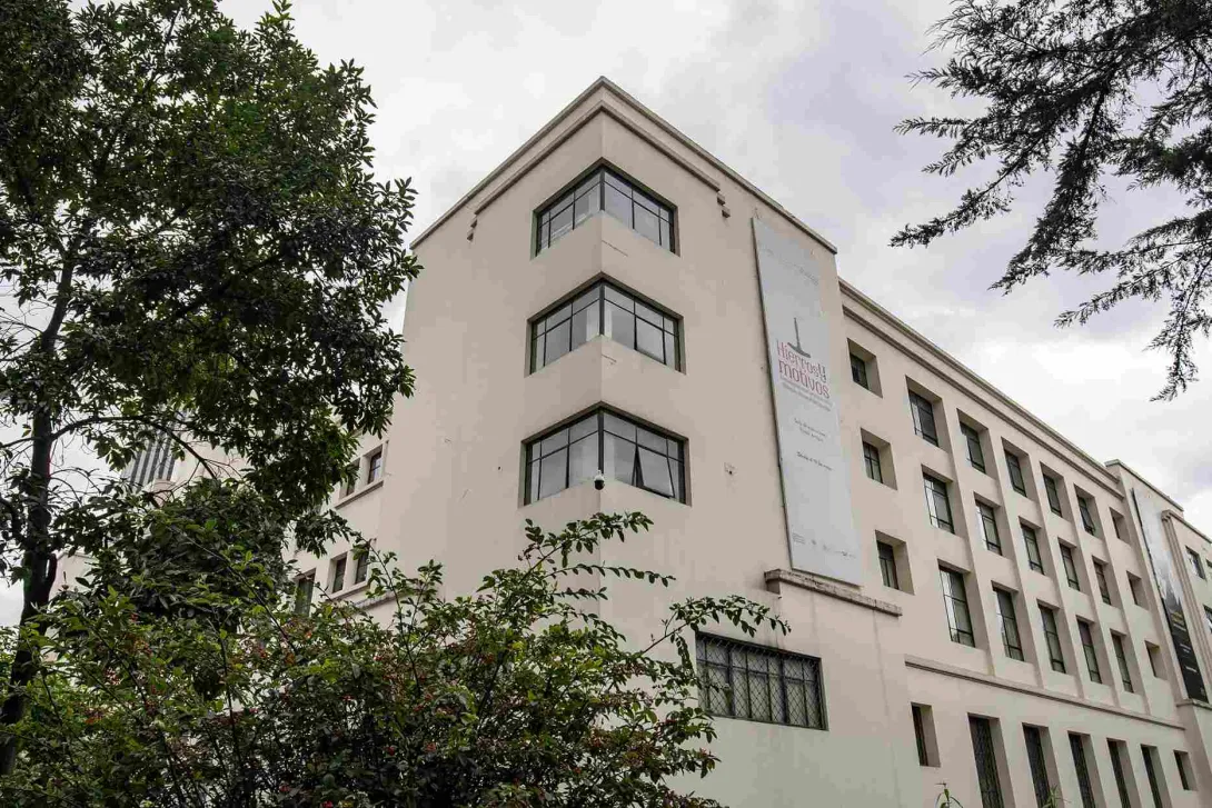 La imagen muestra un edificio de estilo sobrio y elegante, con fachada en tonos claros y grandes ventanas en esquina que aportan modernidad. Está rodeado de árboles y vegetación, lo que le da un ambiente tranquilo.