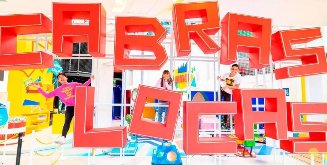 Tres personas posan y juegan entre grandes letras rojas que forman “CABRAS LOCAS” dentro de un espacio recreativo colorido. 