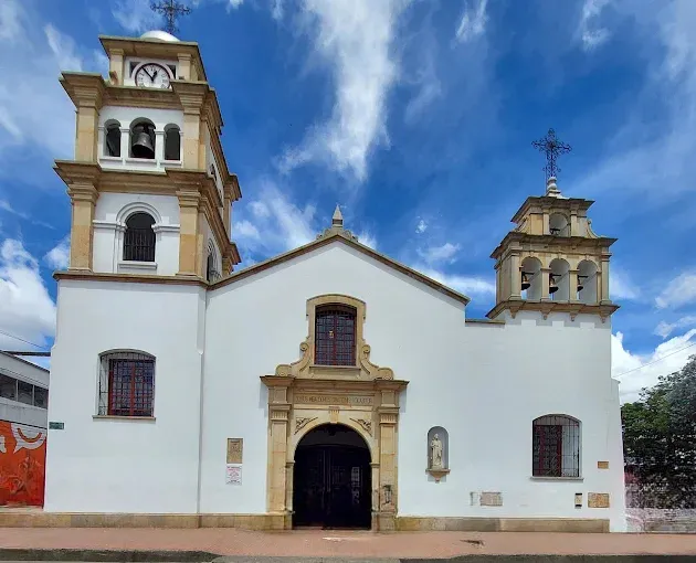 La imagen muestra la fachada de una iglesia de estilo colonial, de paredes blancas y detalles en tonos beige y ocre que resaltan su arquitectura. El edificio se compone de un cuerpo central con una entrada principal enmarcada por un arco ornamentado, sobre el cual se observa una ventana con reja y molduras decorativas. 