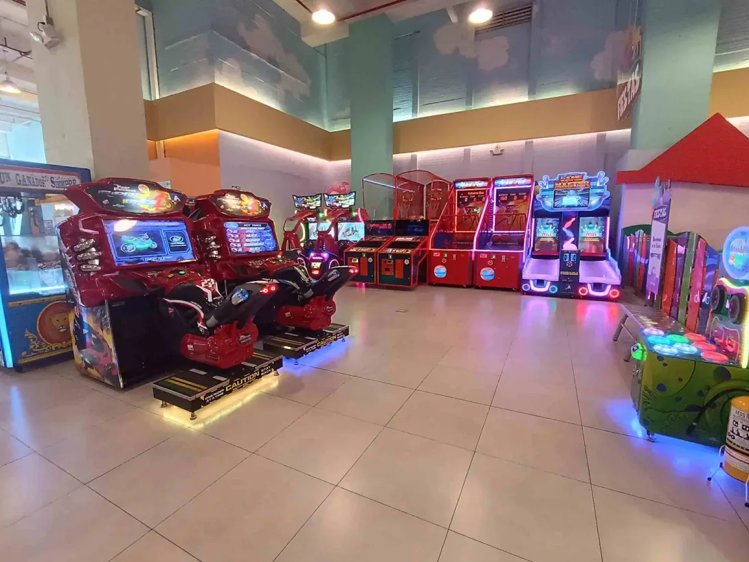 Zona de videojuegos arcade en interior con máquinas de carreras, juegos de canasta y luces de colores.