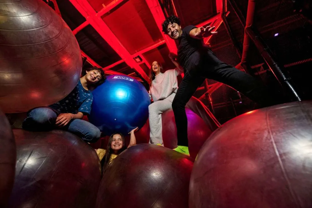 Cuatro personas sonríen y juegan sobre grandes pelotas de ejercicio en un espacio interior iluminado con luces rojas.
