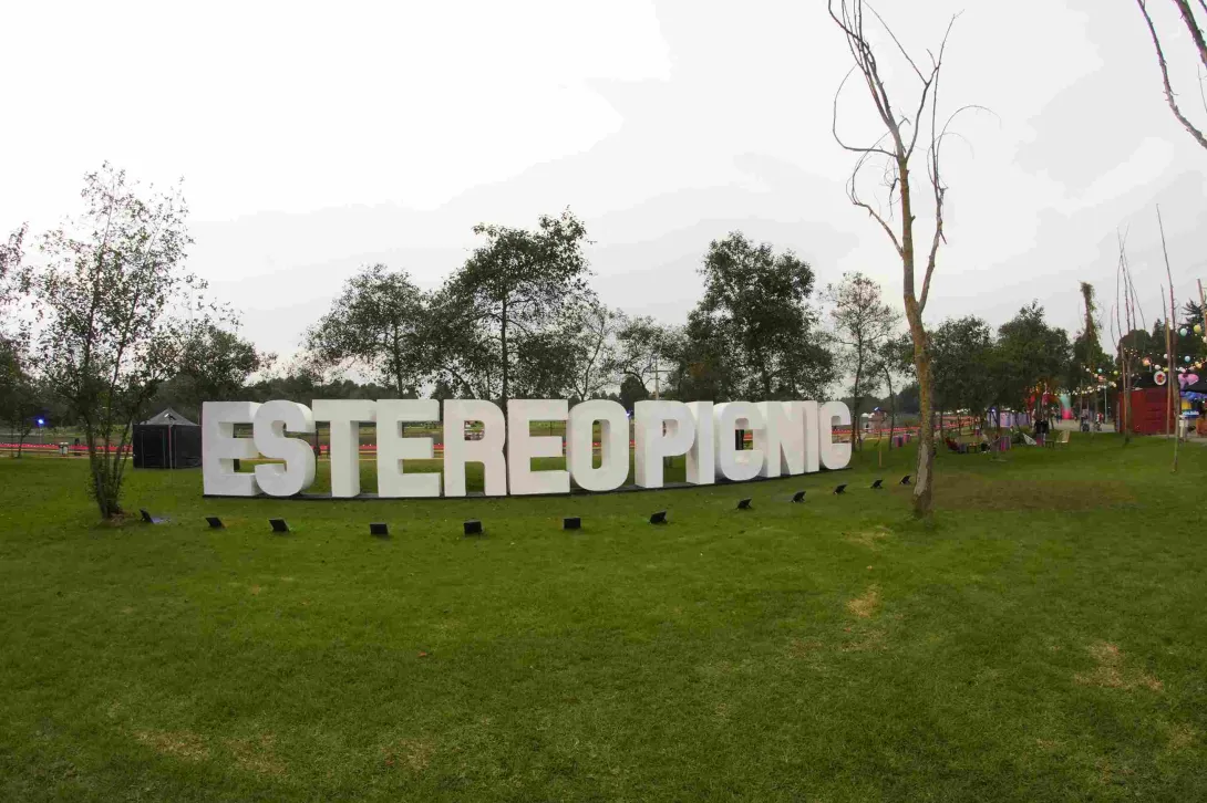 Letras gigantes blancas que forman “ESTÉREO PICNIC” en un parque verde, rodeadas de árboles y estructuras de festival al fondo.