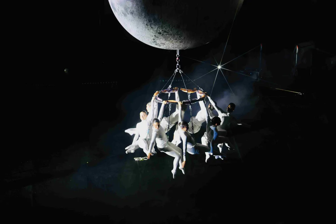 Escena artística en un espacio oscuro: varios acróbatas vestidos de blanco están suspendidos en una estructura colgante bajo una gran luna, iluminados por un foco que resalta la atmósfera teatral y aérea.