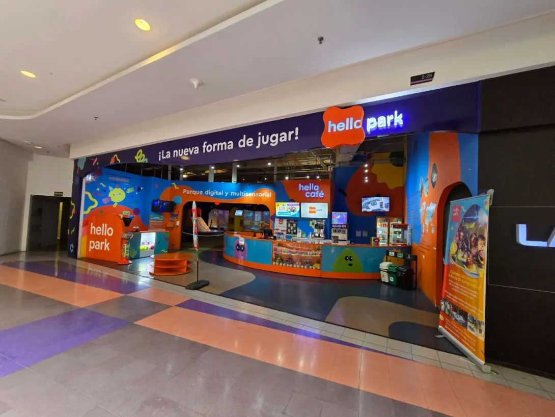 Entrada de “Hello Park” en un centro comercial, con fachada colorida, mostrador de atención, pantallas digitales y letreros que anuncian un parque multisensorial y cafetería.