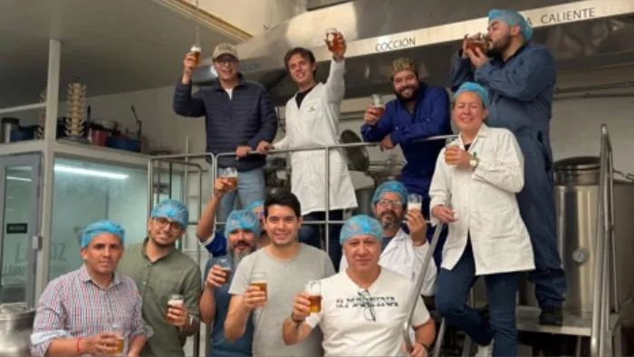 Grupo de once personas en una planta de producción, posando y brindando con vasos de cerveza. La mayoría lleva cofias o gorros de higiene y algunos visten batas blancas, lo que sugiere un entorno de elaboración de alimentos o bebidas.