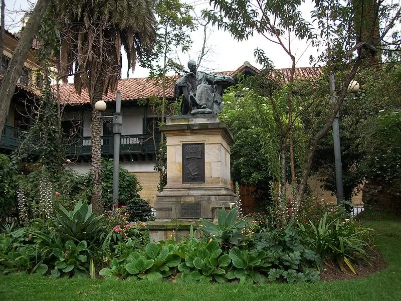 Estatua sobre pedestal en un jardín rodeado de árboles y plantas frente a una casa de estilo tradicional.