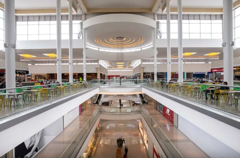 La imagen muestra el interior de un centro comercial amplio y luminoso, con una arquitectura moderna dominada por columnas blancas y grandes ventanales que dejan entrar abundante luz natural. En el nivel superior se observa una zona de comidas con mesas y sillas ocupadas por visitantes, mientras que en el centro se abre un vacío que conecta visualmente con los pisos inferiores mediante escaleras eléctricas.
