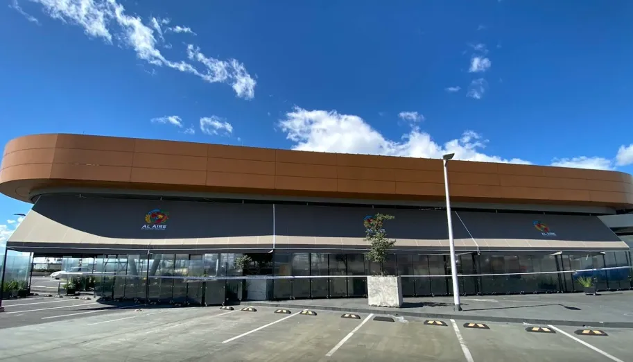 La imagen muestra un moderno edificio comercial de diseño contemporáneo, caracterizado por sus líneas curvas y una fachada elegante en tonos cálidos, que contrastan con el cielo azul intenso salpicado de nubes blancas.