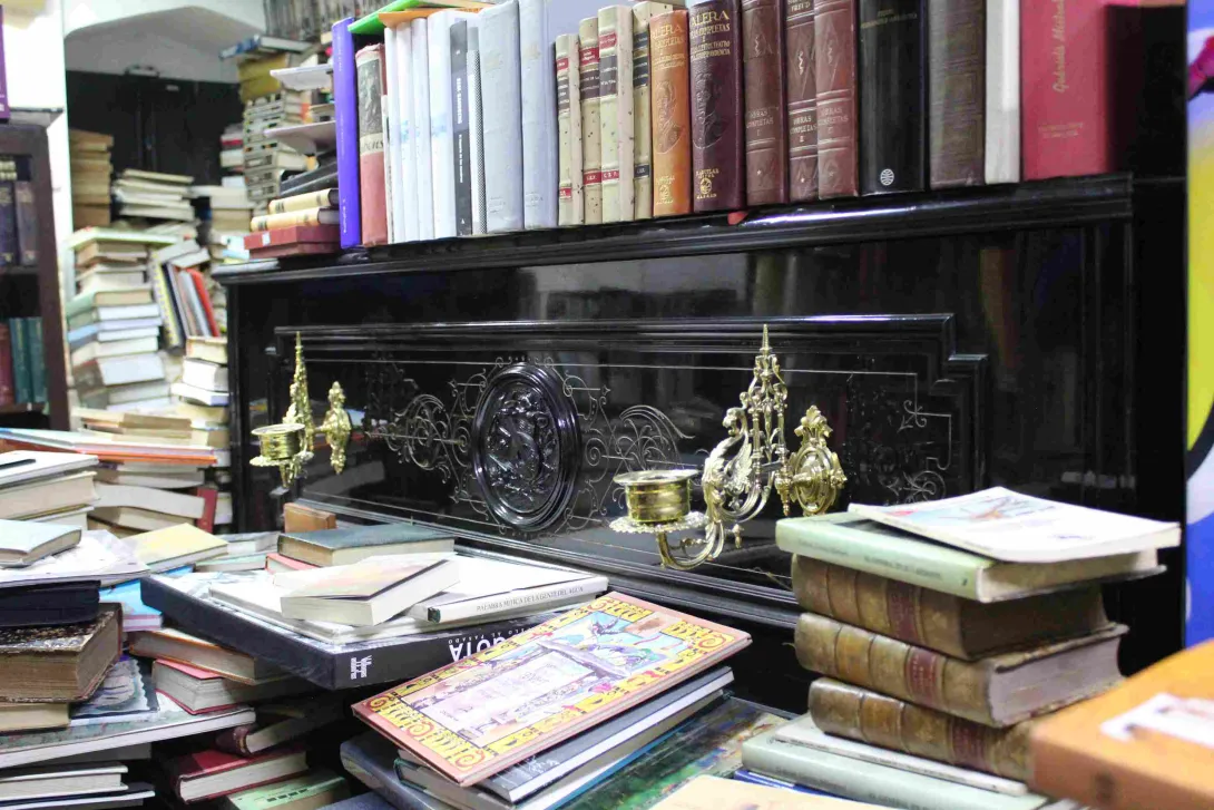 Antique piano with decorative candlesticks, surrounded by stacks of books in a bookstore.