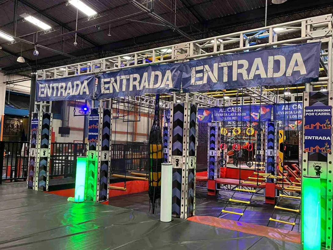 Entrada a un circuito de obstáculos tipo ninja en un parque interior, con estructuras metálicas, cuerdas y luces de colores.