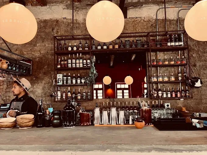Barra de un bar con estilo rústico e industrial, con paredes de ladrillo y estanterías metálicas llenas de botellas de licor y frascos. Sobre la barra de madera se ven utensilios de coctelería, vasos y recipientes. Tres lámparas redondas cuelgan del techo, iluminando el espacio con luz cálida.
