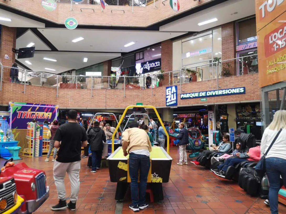  Interior de un centro comercial con zona de juegos, donde varias personas participan en máquinas arcade y mesas de juego, rodeadas de locales y señalización de entretenimiento.
