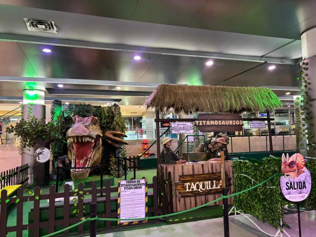  Entrada temática de dinosaurios en un espacio interior, con una gran cabeza de T-Rex, taquilla y señalización de salida