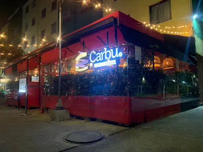 Restaurante con estructura exterior en color rojo ubicado en una esquina urbana durante la noche. El lugar tiene paredes transparentes, iluminación cálida y luces colgantes que crean un ambiente acogedor. En el interior se observan plantas decorativas y mesas ocupadas, mientras en la fachada resalta un letrero iluminado con el nombre “Carbu”.