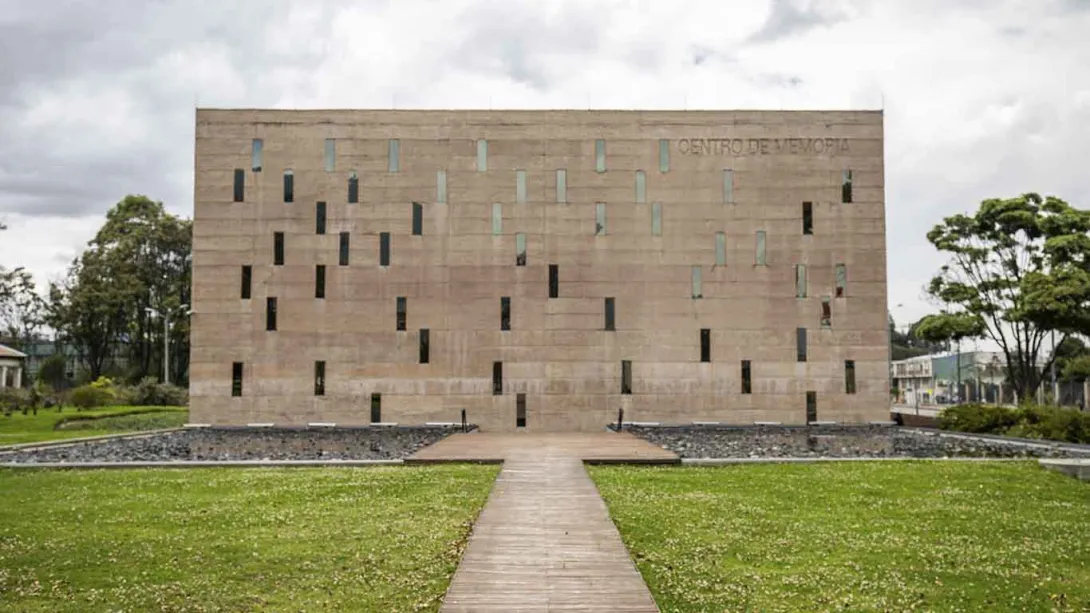La imagen muestra un edificio de arquitectura moderna y minimalista, con una fachada de piedra clara perforada por ventanas verticales irregulares. Un sendero recto conduce hacia la entrada, atravesando una zona verde que rodea la estructura.