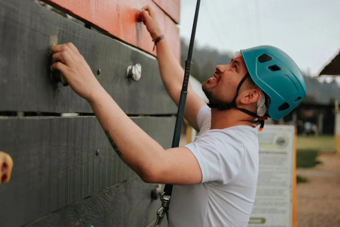 Persona con casco y arnés escala un muro de madera con agarres, en una actividad de aventura al aire libre.