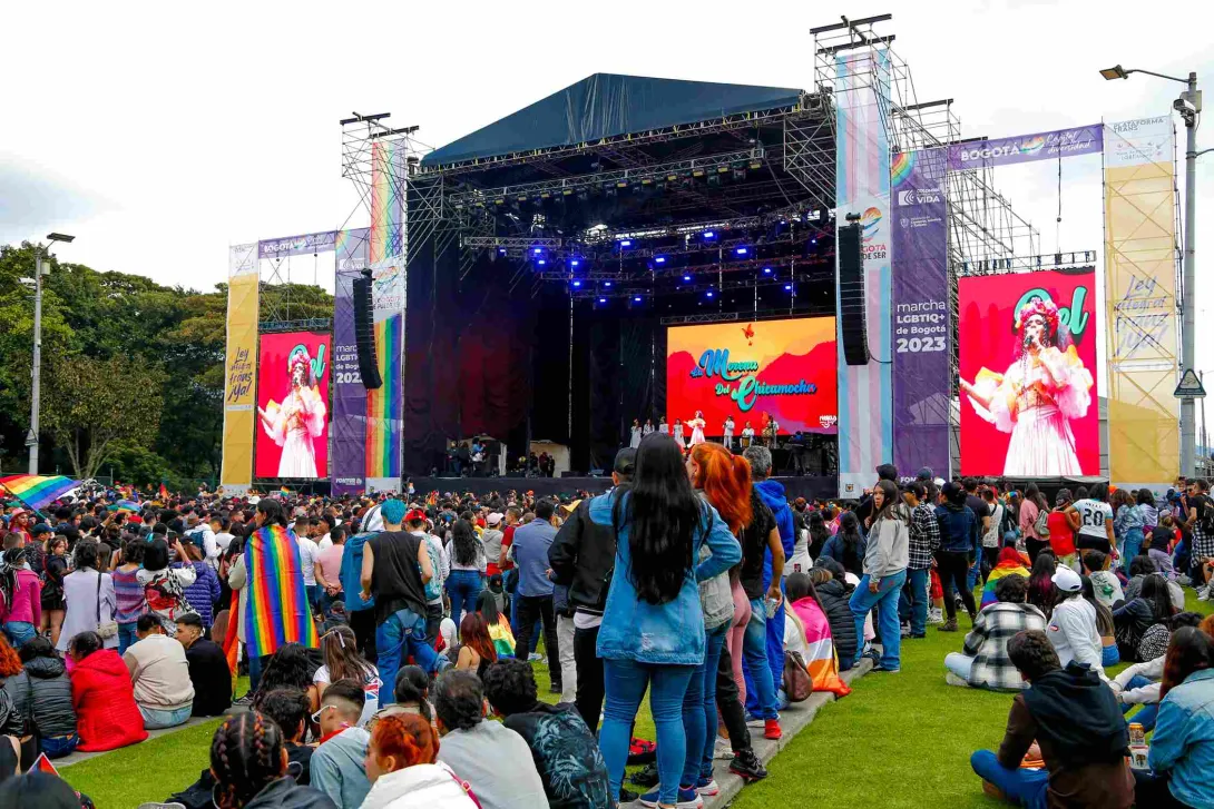 Multitud de personas reunidas frente a un gran escenario al aire libre durante un evento del orgullo LGBTI+ en Bogotá; en las pantallas se ve una artista cantando, mientras asistentes llevan banderas arcoíris y observan el espectáculo sobre el césped.