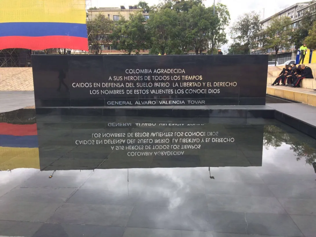  Monumento con placa conmemorativa a héroes caídos, reflejada en una superficie de agua, con bandera de Colombia al fondo.