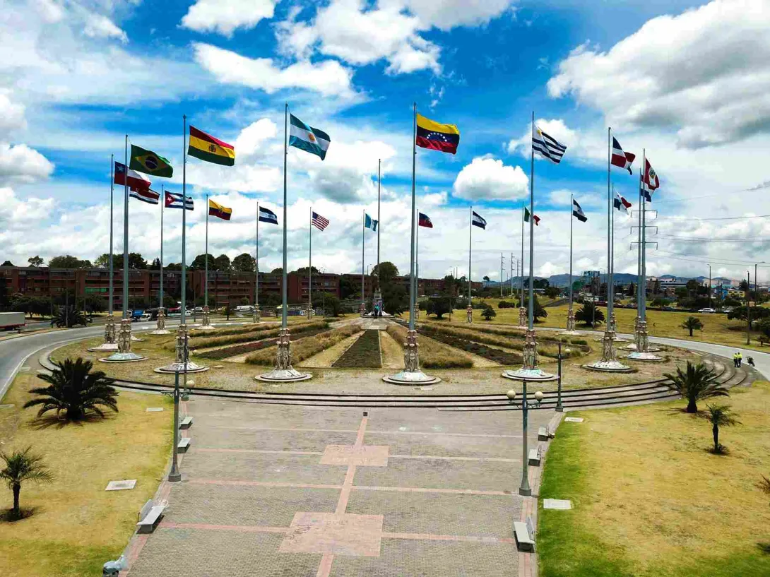  Monumento con múltiples banderas ondeando en una plaza abierta bajo cielo parcialmente nublado.
