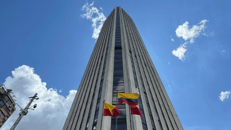 Edificio alto de estilo moderno visto desde abajo, con líneas verticales que acentúan su altura. En la base, varias banderas de Colombia ondean frente a la fachada, bajo un cielo azul con nubes dispersas.