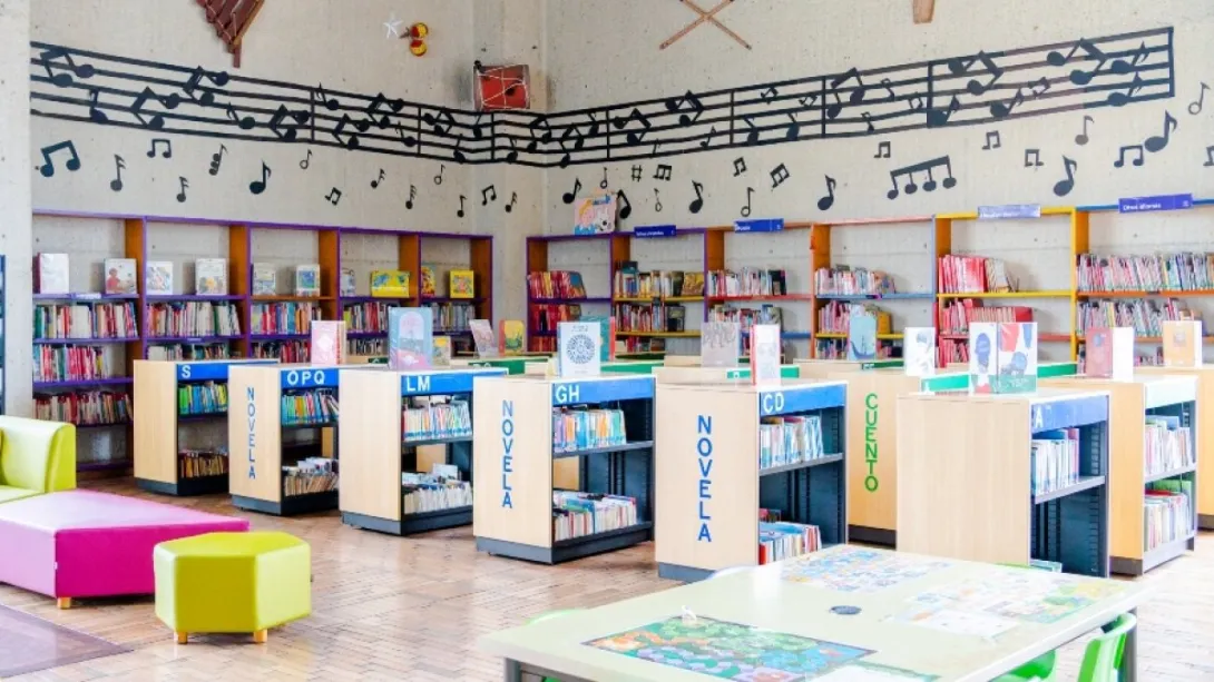 Interior de una biblioteca pública infantil, con estanterías bajas llenas de libros organizados por categorías como “novela” y “cuento”. El espacio es colorido, con muebles en tonos vivos, mesas de lectura y pufs, y una pared decorada con un gran pentagrama y notas musicales que recorre todo el salón, creando un ambiente lúdico y acogedor para niños.