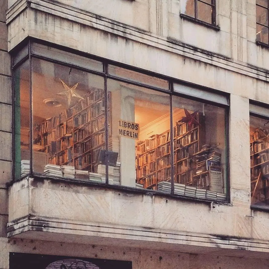 Ventana exterior desde la que se observa la Librería Merlín con estanterías llenas de libros, iluminadas cálidamente desde el interior.