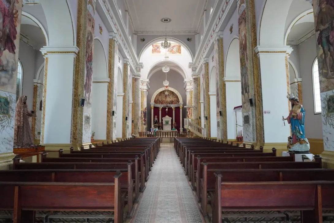 Interior de una iglesia con pasillo central y bancas de madera, columnas decoradas y un altar al fondo enmarcado por arcos y pinturas religiosas.