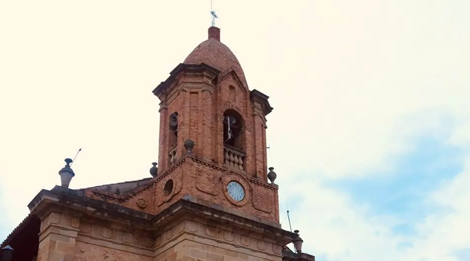 Torre de una iglesia de ladrillo con campanario y reloj, coronada por una cruz, contra un cielo claro con nubes.