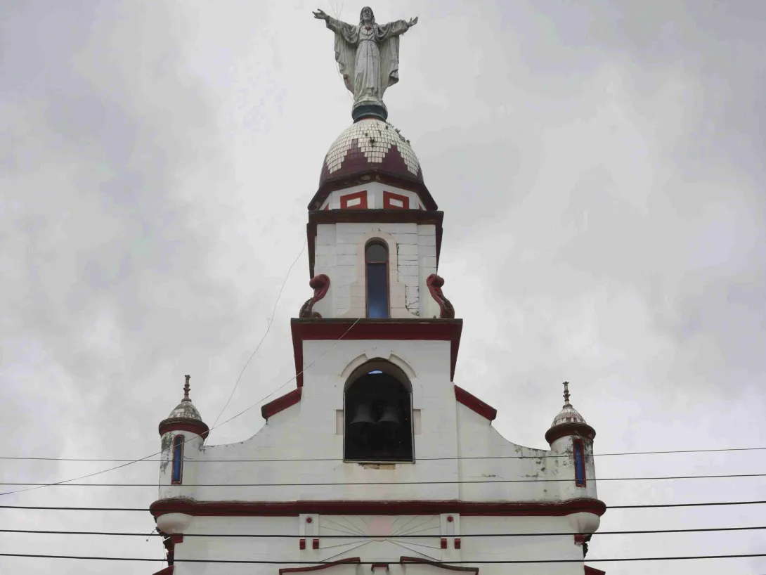  Torre de iglesia blanca con detalles rojos, coronada por una estatua de Cristo con los brazos abiertos, contra un cielo nublado.