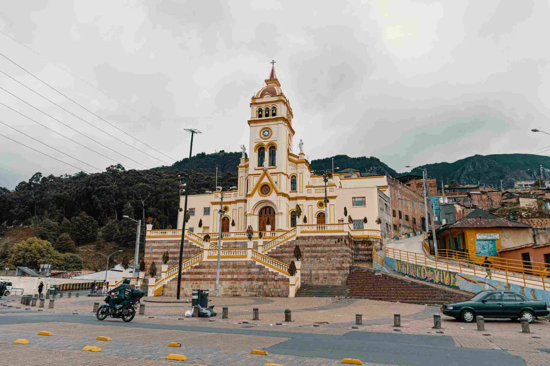  Plano general de la fachada de una iglesia color marrón claro, tras la cual hay montañas. Al frente hay una calle con un automovil estacionado y un motociclista.