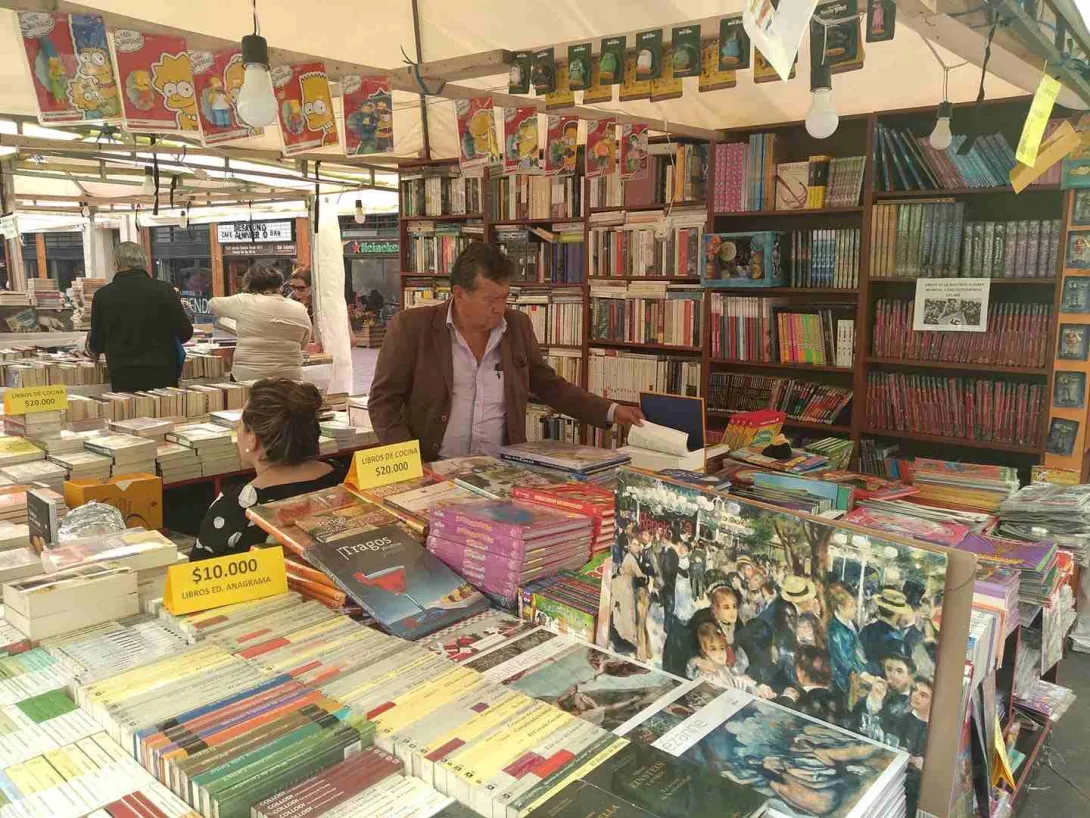Puesto de venta de libros en un mercado, con estanterías llenas y personas explorando y comprando entre títulos variados.