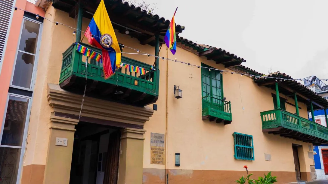 Fachada de casa colonial con balcones de madera pintados de verde, decorada con banderas de Colombia y colores del orgullo, en una calle de estilo tradicional.