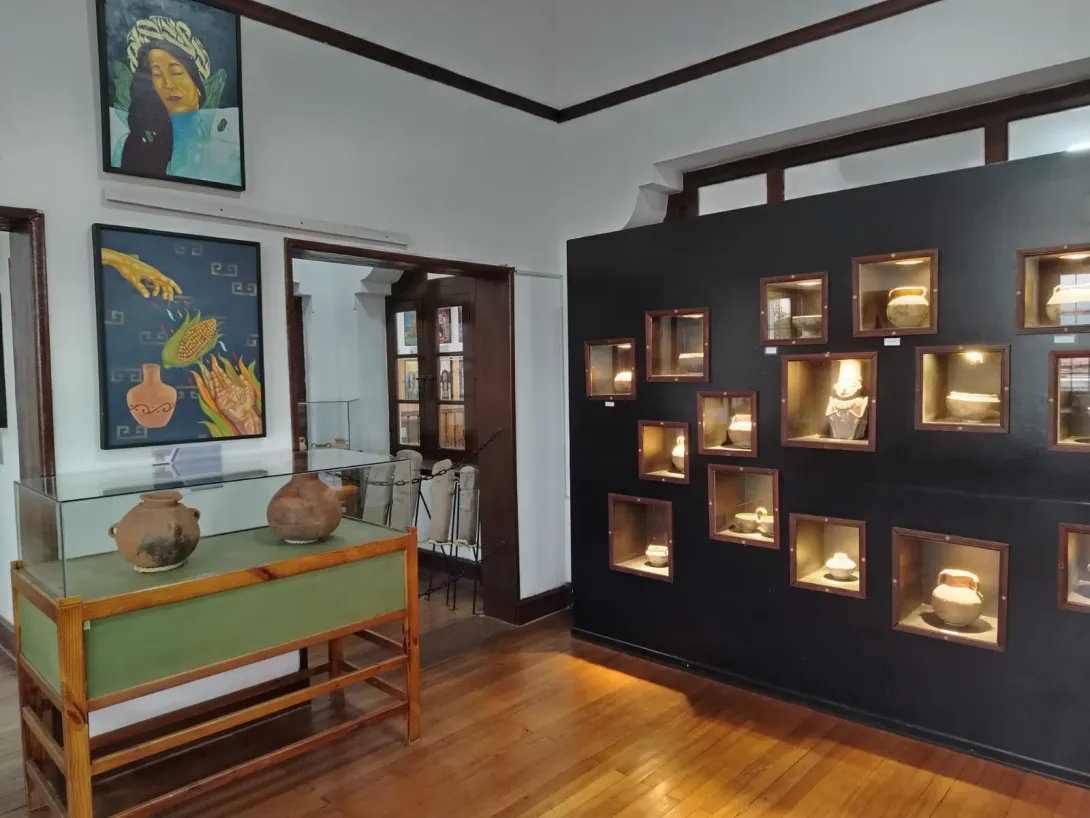  Sala de exposición de un museo arqueológico con piso de madera, vitrinas de vidrio y nichos iluminados en la pared que exhiben piezas de cerámica precolombina. En las paredes se observan pinturas de temática indígena y, al fondo, una puerta comunica con otra sala del museo.