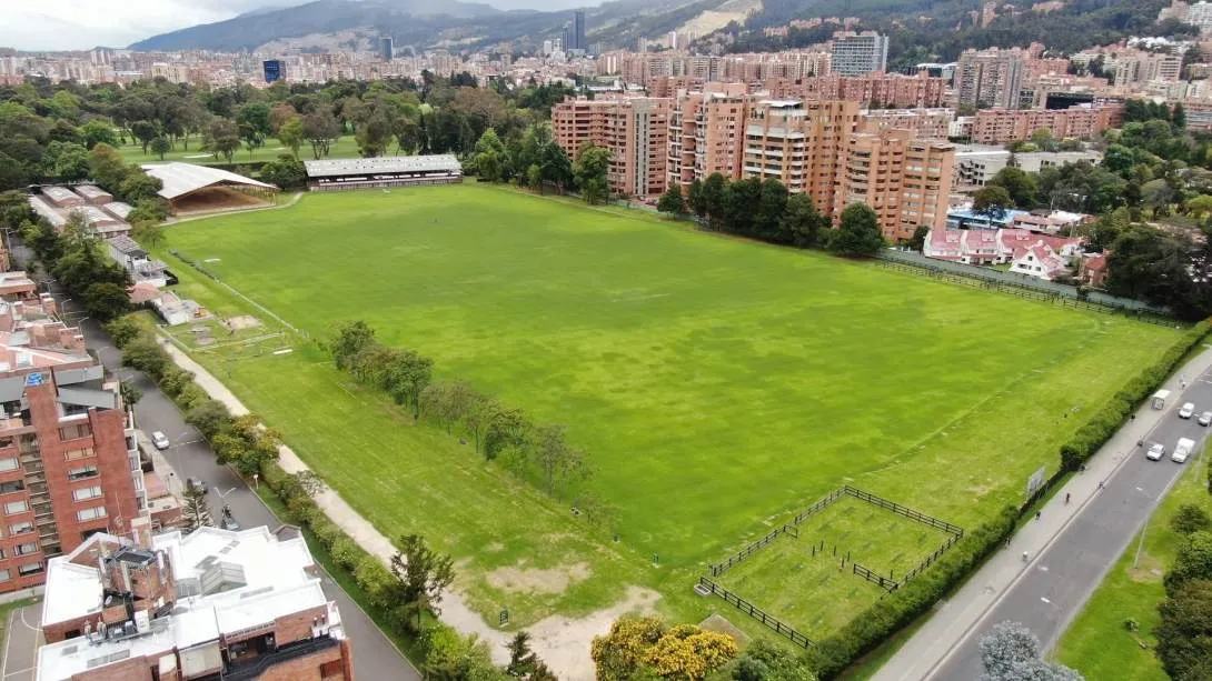 Aerial view of a large grass field surrounded by trees, residential buildings, and urban roads, with the eastern hills in the background.