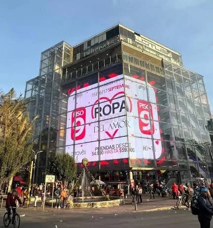 Vista exterior de un moderno edificio comercial de varios pisos con fachada de vidrio y una gran pantalla publicitaria digital. En el primer plano se observa una concurrida intersección con peatones, ciclistas y vehículos, reflejando el dinamismo del sector comercial de San Victorino, en Bogotá.