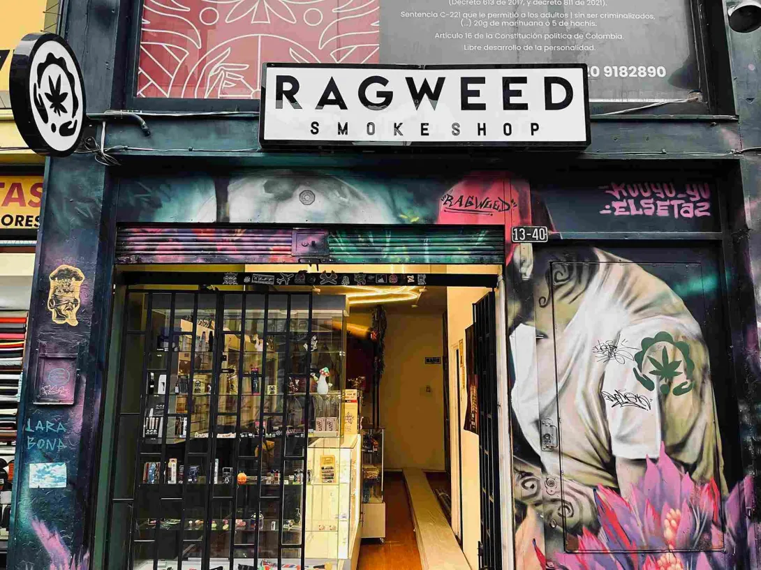 Fachada de una tienda con el letrero "Ragweed Smokeshop", rodeada de murales de arte urbano y grafitis. La entrada, con reja negra y vitrinas iluminadas, exhibe diversos productos en su interior.
