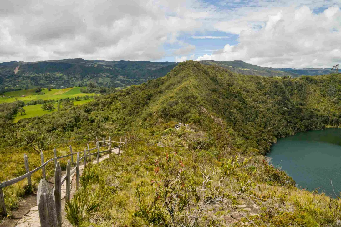 Sendero ecológico con barandas de madera entre montañas andinas, junto a una laguna de aguas verdes y un paisaje de vegetación nativa bajo un cielo parcialmente nublado.