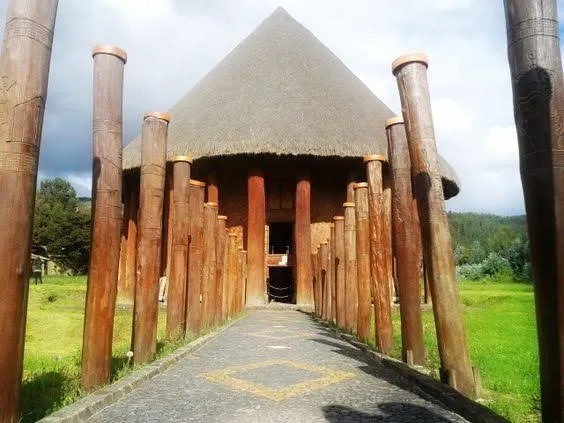 Camino de piedra flanqueado por altos postes de madera que conduce a un templo circular con techo de paja, rodeado de zonas verdes bajo un cielo parcialmente nublado.