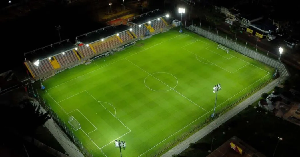 Vista aérea nocturna del Estadio Olaya Herrera con la cancha de fútbol iluminada, graderías a un costado y reflectores encendidos alrededor del escenario deportivo.
