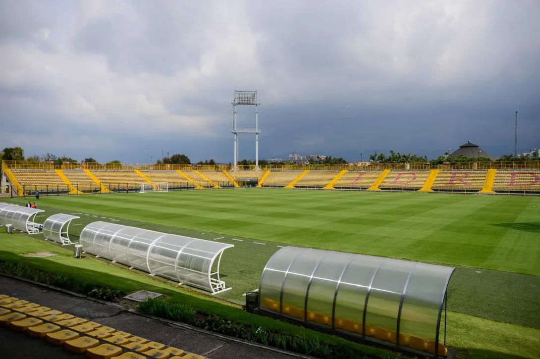  Vista panorámica del Estadio de Techo con la cancha de césped, graderías amarillas y los bancos de suplentes en primer plano bajo un cielo nublado.