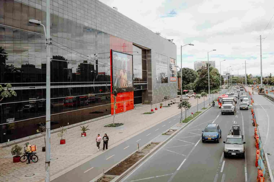 Una amplia avenida bordea la moderna fachada del Centro Comercial Metrópolis, donde el movimiento de vehículos, peatones y ciclistas refleja la dinámica cotidiana de este sector de Bogotá.