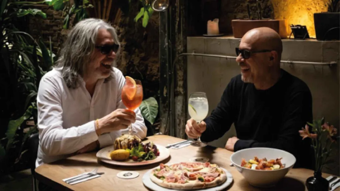  Dos hombres con gafas oscuras sonríen y brindan con cócteles frente a una mesa llena de comida en un restaurante.