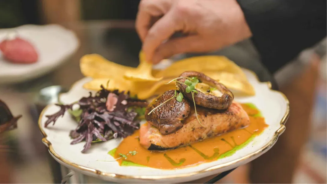 Mano sirviendo un plato de salmón en salsa con champiñones fritos, hojas verdes y crujientes de plátano encima.